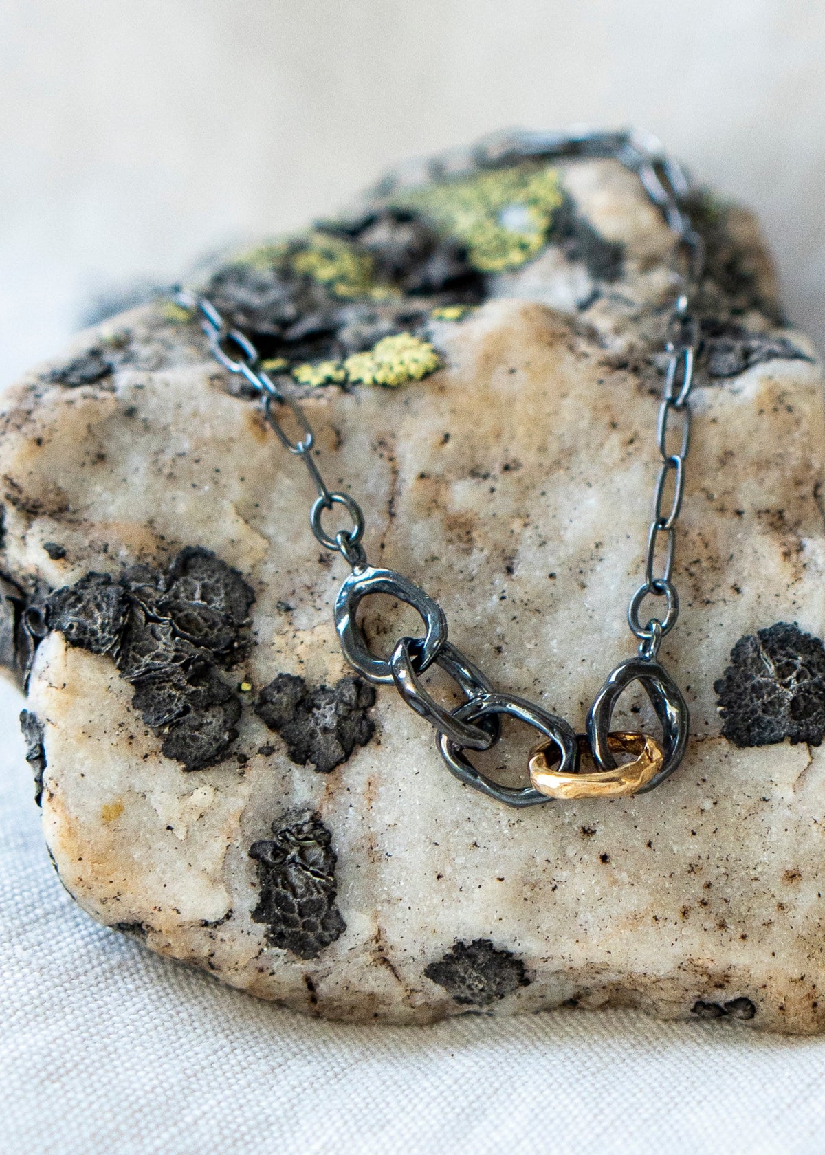 A close-up of a chain necklace with dark oxidized metal links and one polished gold link, displayed on a textured stone surface.