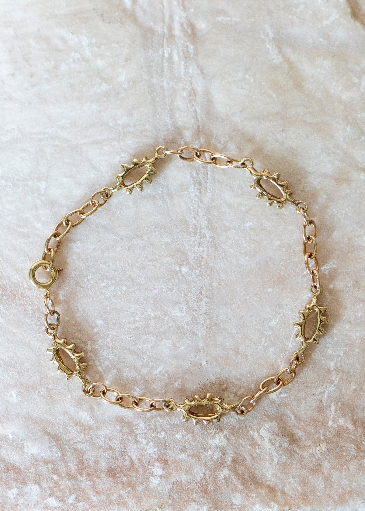A gold chain bracelet with oval links and sunburst-shaped accents, displayed on a textured beige fabric background.