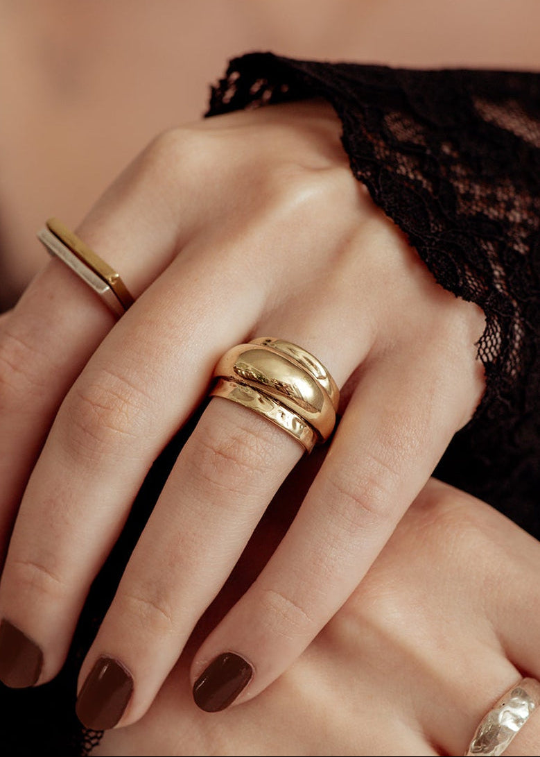 A close-up of a hand wearing multiple gold rings, including a wide polished band, a textured engraved band, and a slim rectangular band, with dark brown nail polish and a black lace sleeve visible.