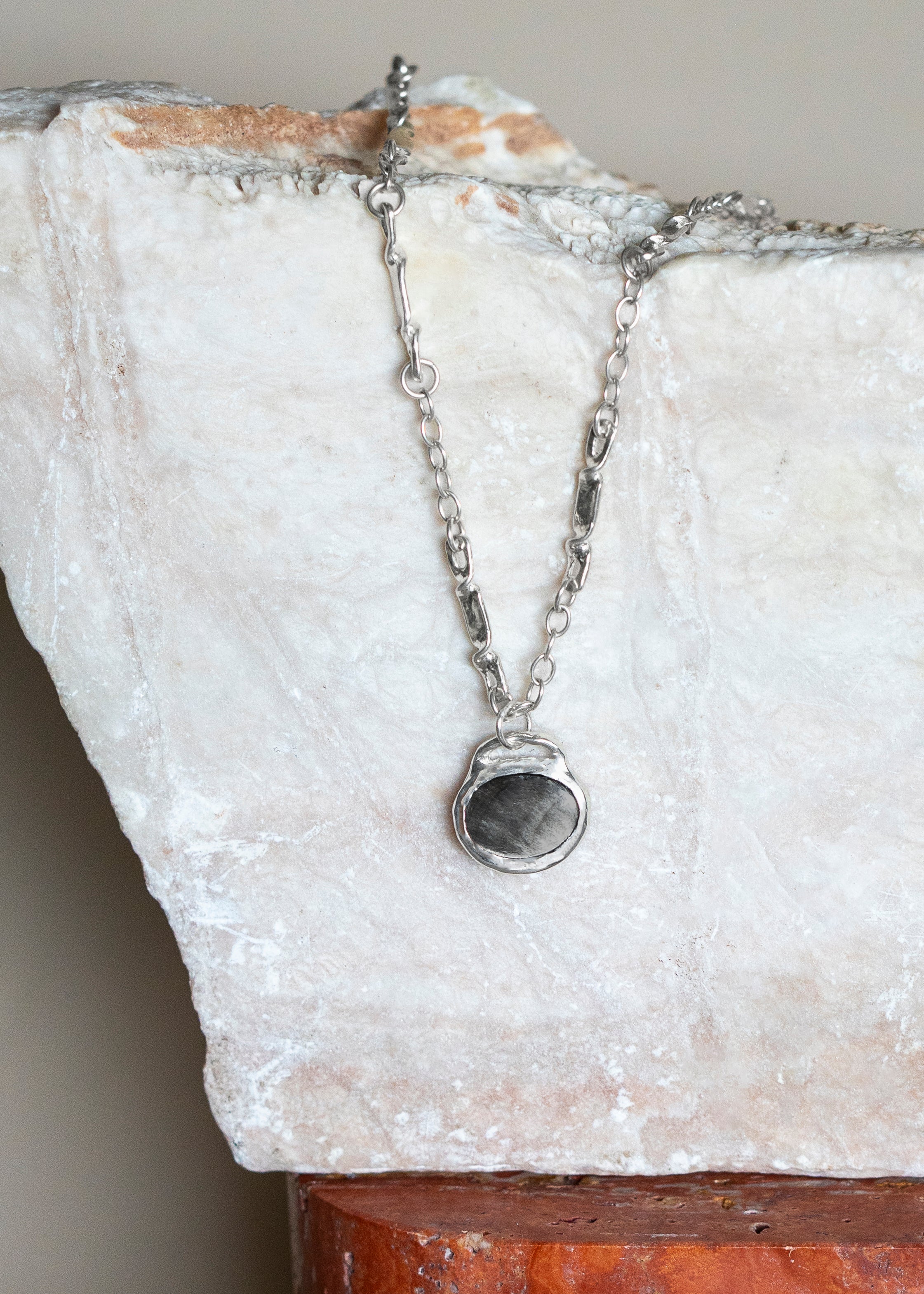 A silver MINA chain necklace with  an oval Black. other of Pearl textured pendant draped over a white stone display block.